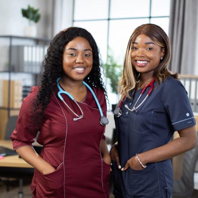 Close up shot of two female African American doctors or nurses in colorful medical scrubs, smiling while standing in modern hospital room. Two cheerful afro women healthcare practitioners