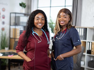 Close up shot of two female African American doctors or nurses in colorful medical scrubs, smiling while standing in modern hospital room. Two cheerful afro women healthcare practitioners